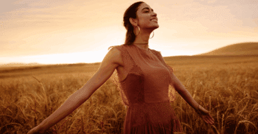 Uma mulher jovem está em um campo durante o pôr do sol. Ela levanta o rosto e sorri sentindo a brisa do vento. Ela está com os dois braços abertos.