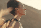 Uma mulher jovem está fazendo uma trilha em um ambiente de natureza. Ela sente a luz do pôr do sol no rosto, fecha os olhos e sorri.