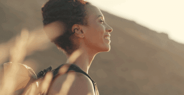 Uma mulher jovem está fazendo uma trilha em um ambiente de natureza. Ela sente a luz do pôr do sol no rosto, fecha os olhos e sorri.