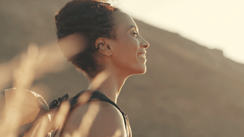 Uma mulher jovem está fazendo uma trilha em um ambiente de natureza. Ela sente a luz do pôr do sol no rosto, fecha os olhos e sorri.