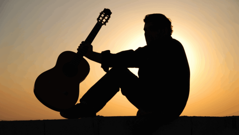 A silhueta de um homem sentado em uma mureta segurando seu violão está no foco da imagem. Atrás dele, há a luz do pôr do sol.