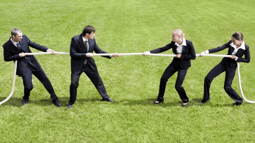 Uma dupla de homens está puxando uma corda de um lado e uma dupla de mulheres está puxando a mesma corda do outro lado. Todos se vestem com roupas sociais, indicando a competição no trabalho.