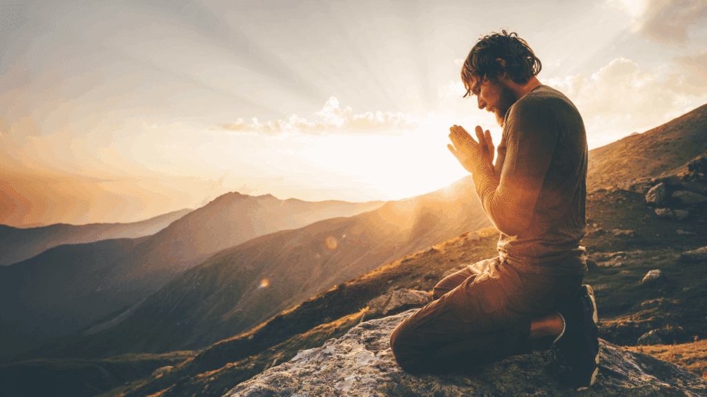 Um homem está de joelhos e com as mãos unidas em forma de oração. Ele está em uma montanha. Ao fundo, a luz do pôr do sol reflete atrás da montanha.