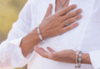 A imagem foca nas mãos de uma mulher. Ela coloca as suas mãos na altura do peito, indicando gratidão, paz. Ela veste roupas brancas e usa pulseiras e um colar.