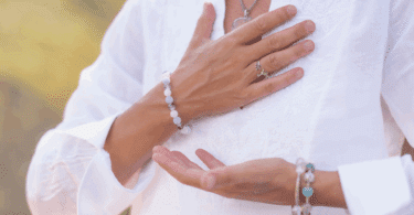 A imagem foca nas mãos de uma mulher. Ela coloca as suas mãos na altura do peito, indicando gratidão, paz. Ela veste roupas brancas e usa pulseiras e um colar.
