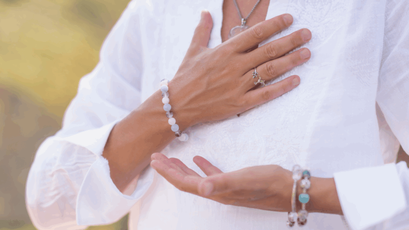 A imagem foca nas mãos de uma mulher. Ela coloca as suas mãos na altura do peito, indicando gratidão, paz. Ela veste roupas brancas e usa pulseiras e um colar.