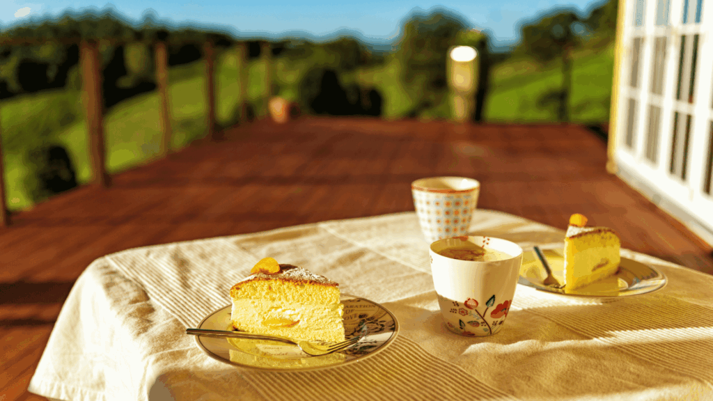 Uma mesa de café da tarde em que há dois pedaços de bolo e duas xícaras de café está em destaque na imagem. A mesa está em uma varanda. Ao fundo, há o céu limpo, as montanhas e a luz do pôr do sol.