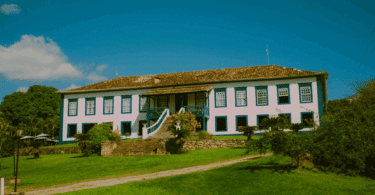 Uma casa colonial grande está em destaque na imagem. Na frente da casa, há um gramado e outras plantas. Ao fundo, há o céu limpo.
