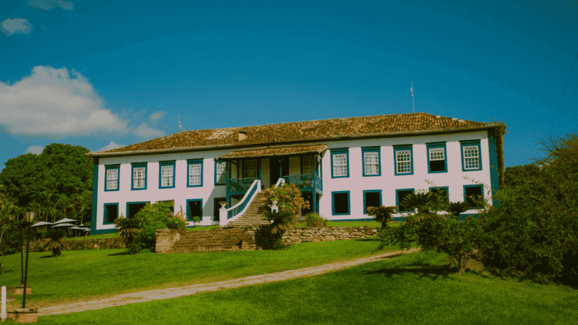 Uma casa colonial grande está em destaque na imagem. Na frente da casa, há um gramado e outras plantas. Ao fundo, há o céu limpo.