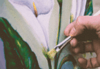 A mão de um pintor que está pintando o desenho de uma flor branca em um quadro está em destaque na imagem.