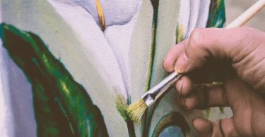 A mão de um pintor que está pintando o desenho de uma flor branca em um quadro está em destaque na imagem.