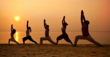 Imagem de cinco pessoas, três mulheres e dois homens praticando yoga ou ioga na praia, ao lado do pôr do Sol.