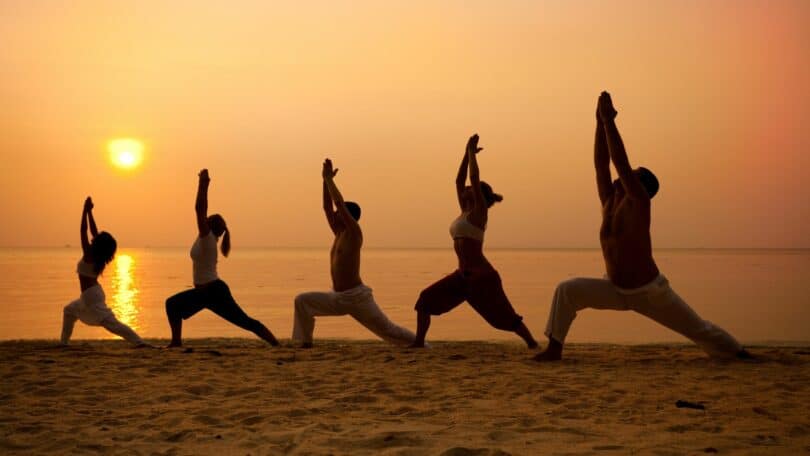 Imagem de cinco pessoas, três mulheres e dois homens praticando yoga ou ioga na praia, ao lado do pôr do Sol.