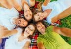 Imagem de 5 pessoas deitadas em um gramado sobre uma manta xadrez. Elas estão fazendo uma selfie de todos felizes, trazendo o conceito de pessoas com energia positiva.