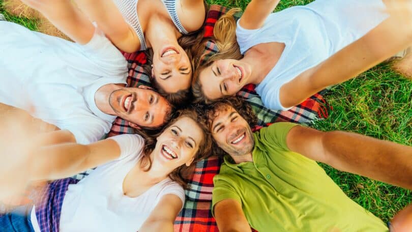 Imagem de 5 pessoas deitadas em um gramado sobre uma manta xadrez. Elas estão fazendo uma selfie de todos felizes, trazendo o conceito de pessoas com energia positiva.