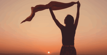 No foco da imagem, há uma mulher levantando um lenço ao vento. Ela contempla o pôr do sol.