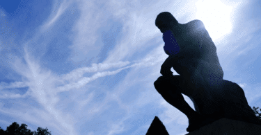 Na imagem, há a silhueta da estátua que representa "O Pensador" de Auguste Rodin. Ao fundo, há o céu azul e a luz do sol.