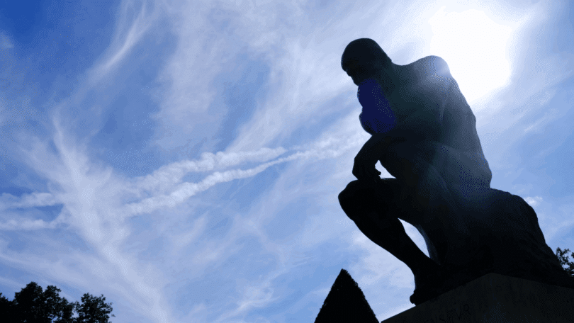 Na imagem, há a silhueta da estátua que representa "O Pensador" de Auguste Rodin. Ao fundo, há o céu azul e a luz do sol.