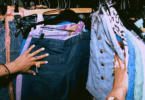 No foco da imagem, há as mãos de uma mulher escolhendo roupas em uma loja vintage.