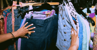 No foco da imagem, há as mãos de uma mulher escolhendo roupas em uma loja vintage.