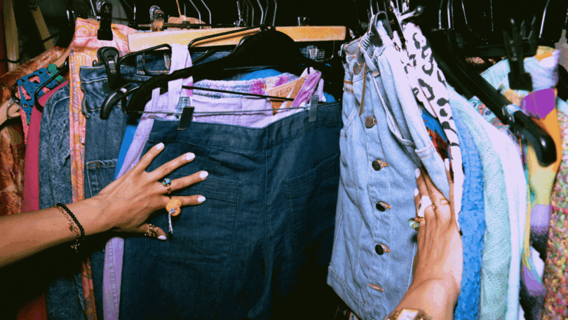 No foco da imagem, há as mãos de uma mulher escolhendo roupas em uma loja vintage.
