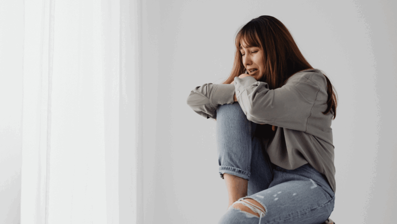 No foco da imagem, há uma jovem mulher sentada e chorando.