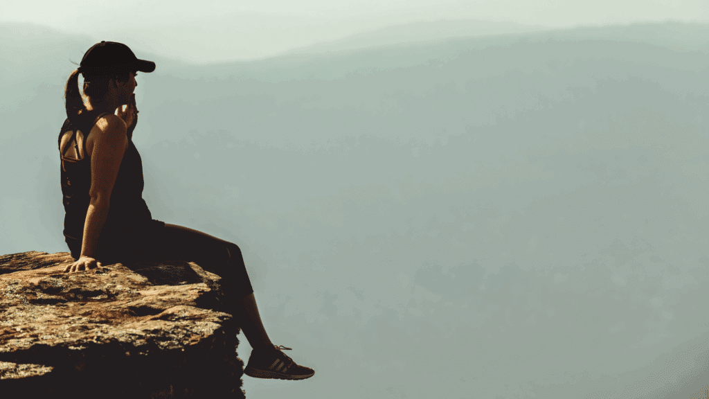 Um mulher sentada na beira de uma montanha ou penhasco está em destaque na imagem. Ela contempla a paisagem.