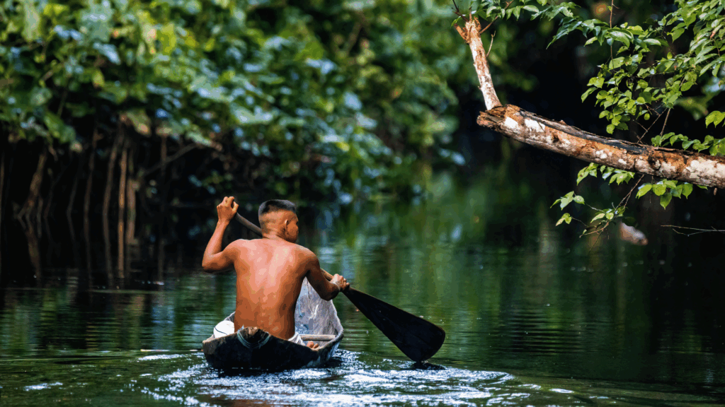 No foco da imagem, há um homem indígena navegando em seu barco de madeira em um rio da Amazônia.
