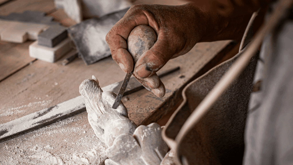 No foco da imagem, há a mão de um artesão esculpindo uma escultura de uma mulher em uma pedra.