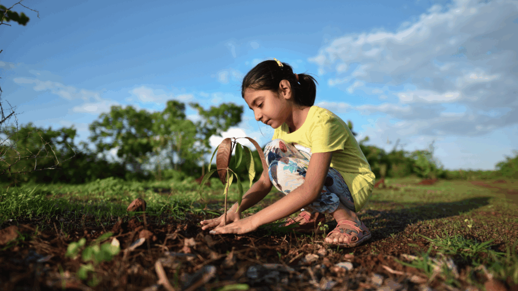 No foco da imagem, há uma menina plantando uma árvore. Ao fundo, há um céu azul com poucas nuvens.