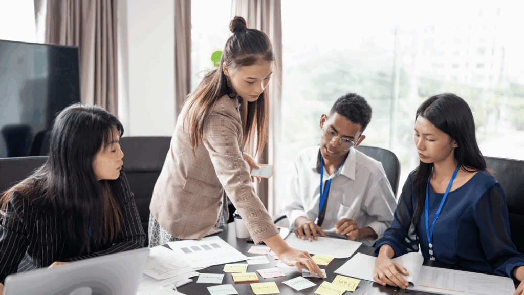Um grupo de funcionários de uma empresa está trabalhando em um projeto. Em destaque e em pé, uma mulher jovem lidera.