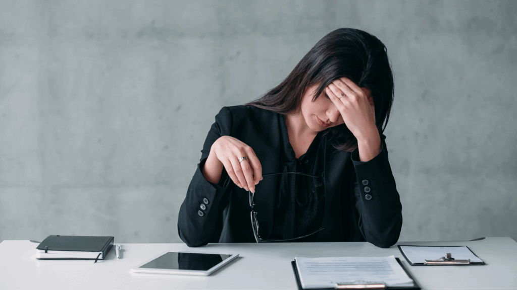 Na imagem, um mulher de negócios está cabisbaixa e com uma mão na testa, indicando estar estressada ou sobrecarregada.