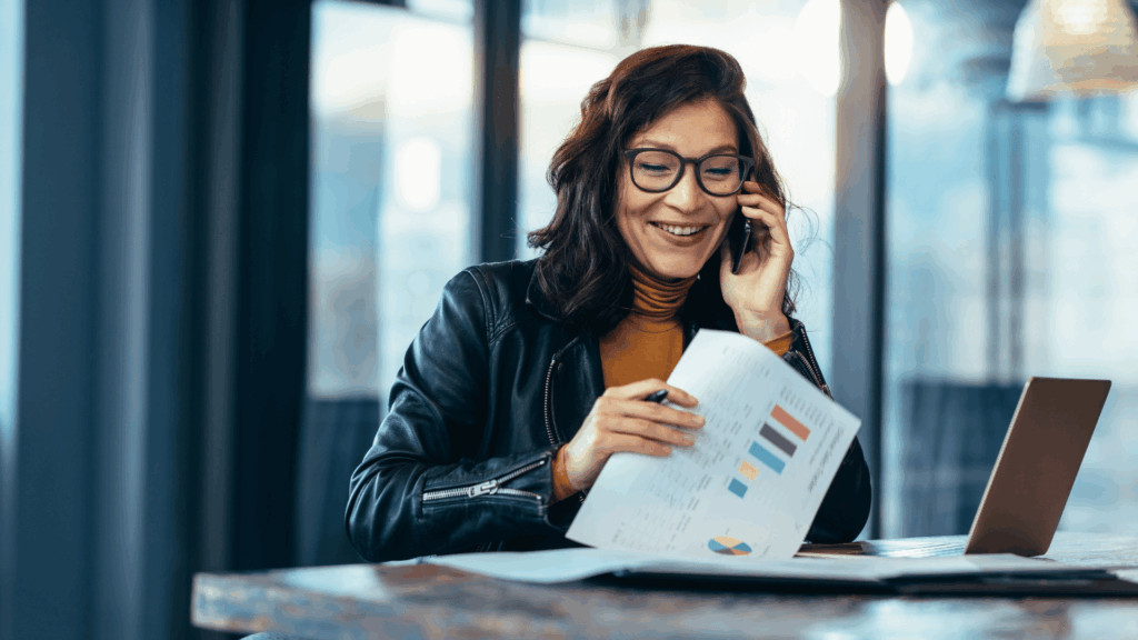 Na imagem, uma mulher de negócios está no seu ambiente de trabalho falando ao celular. Ela segura alguns relatórios e sorri.