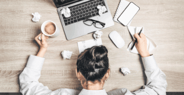 Na imagem, uma mulher de negócios está cabisbaixa, indicando estar sobrecarregada ou cansada. A sua mesa de trabalho está desorganizada e com alguns papéis descartados.