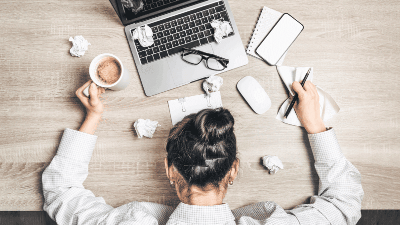 Na imagem, uma mulher de negócios está cabisbaixa, indicando estar sobrecarregada ou cansada. A sua mesa de trabalho está desorganizada e com alguns papéis descartados.
