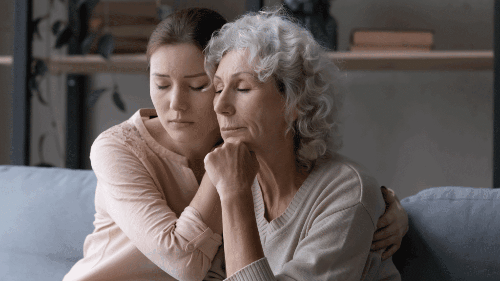 Uma mãe idosa e uma filha adulta estão sentadas em um sofá. As duas apresentam uma feição de tristeza no rosto e ambas se consolam.