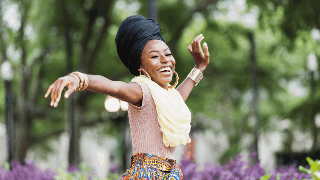 Na imagem, há uma jovem mulher negra. Ela está dançando e sorrindo. Ela usa um turbante, roupas coloridas e acessórios grandes.