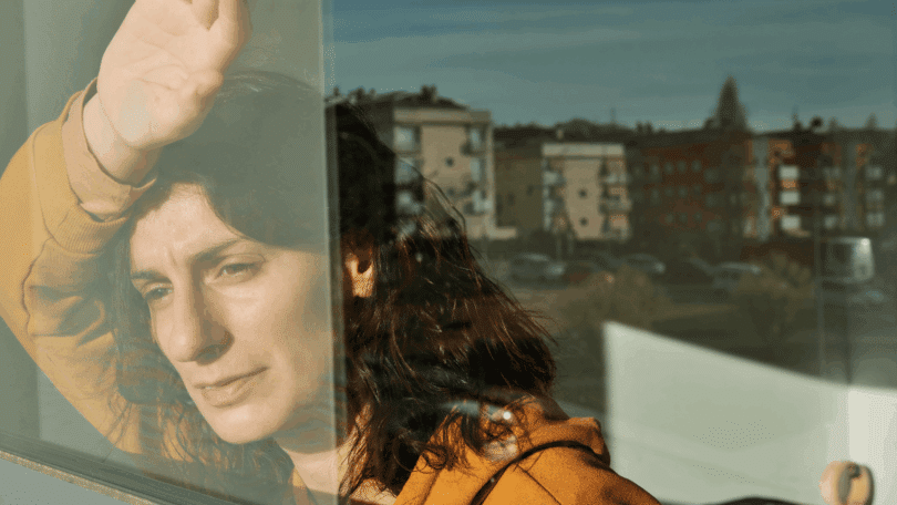 Na imagem, há uma mulher olhando pela janela. Ela está refletindo ou contemplando algo.