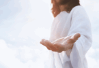 Na imagem, Jesus está estendendo uma de suas mãos para frente. Ao fundo, há nuvens e luz.
