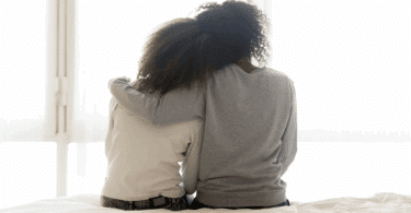Na imagem, há uma mãe abraçando sua filha adolescente, indicando apoio. Elas estão sentadas em uma cama e de costas.
