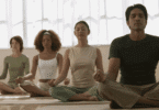 Grupo de pessoas meditando em posição de lótus, com olhos fechados e expressão serena.