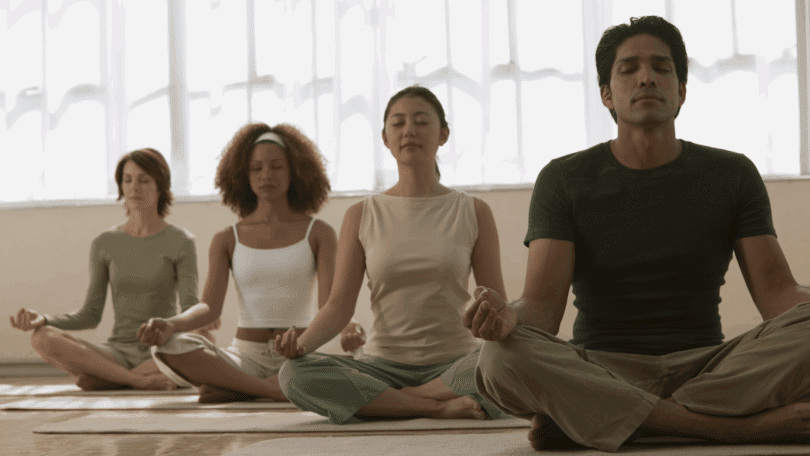 Grupo de pessoas meditando em posição de lótus, com olhos fechados e expressão serena.