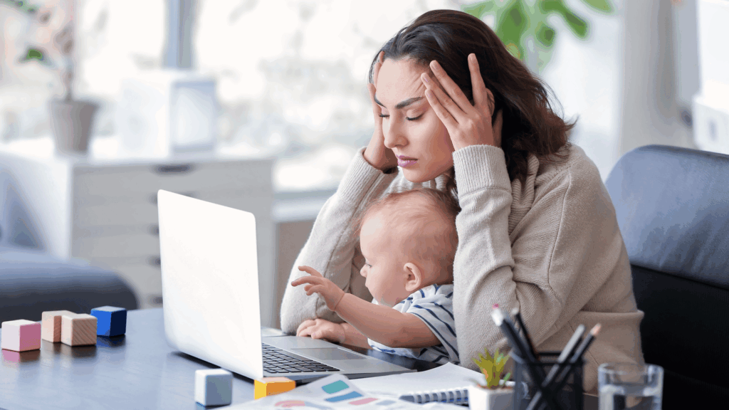 Na imagem, há uma mulher fazendo home office. Ela coloca as mãos na testa, segura seu filho e está cansada.