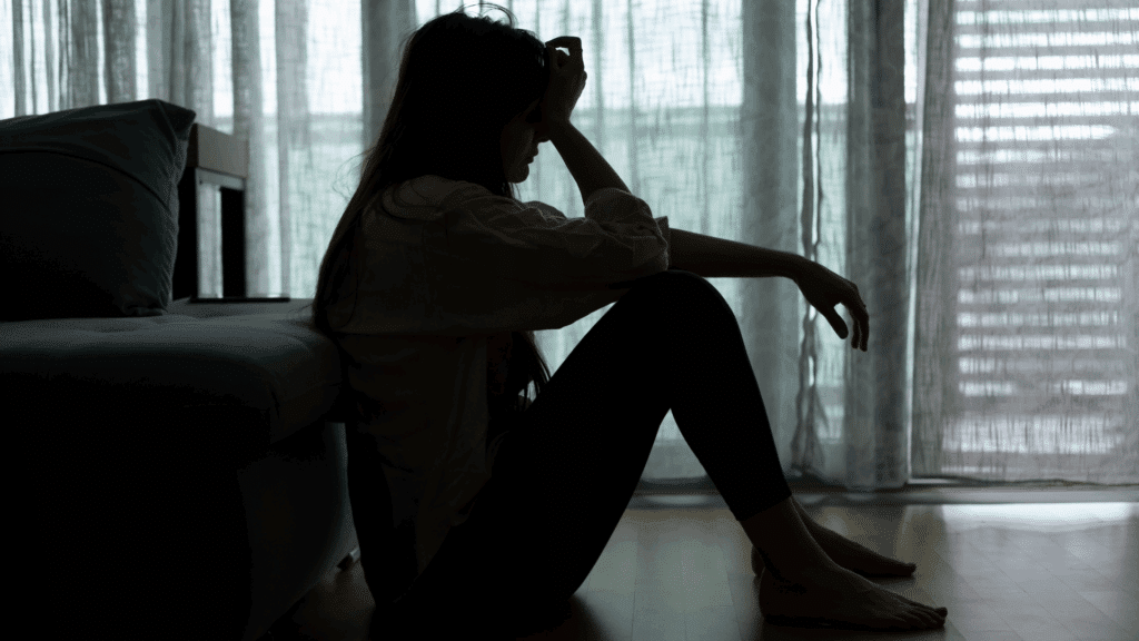 Na imagem, há uma mulher sentada no chão do quarto. Ela coloca uma mão na testa, indicando estar triste ou abalada.