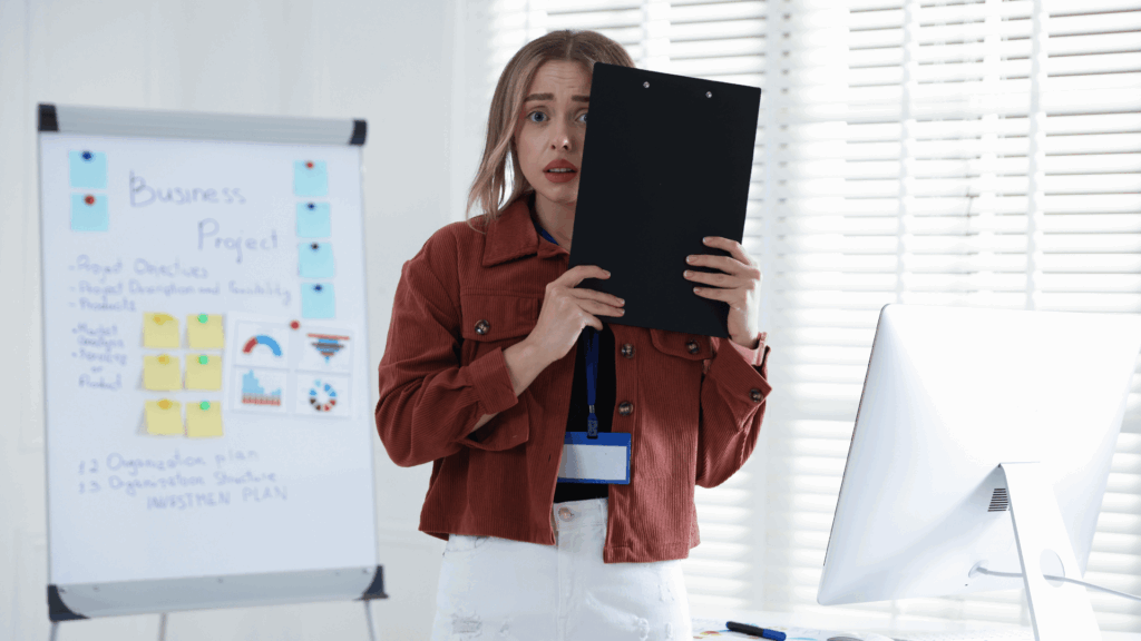 Na imagem, uma mulher de negócios vai apresentar seu projeto. Ela está com medo, tem a expressão de estar assustada e segura uma prancheta nas mãos.