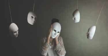 Imagem de uma mulher colocando um máscara sobre o rosto. Várias outras máscaras compõem a imagem, ficando penduradas, simbolizando o velho ditado que diz: o que as pessoas dizem sobre você revela mais sobre elas do que sobre você.