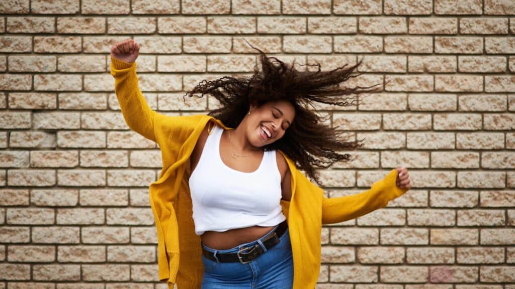 Imagem de fundo de uma parede de tijolos bege claro e em destaque uma mulher negra sorrindo e feliz, simbolizando a autenticidade.