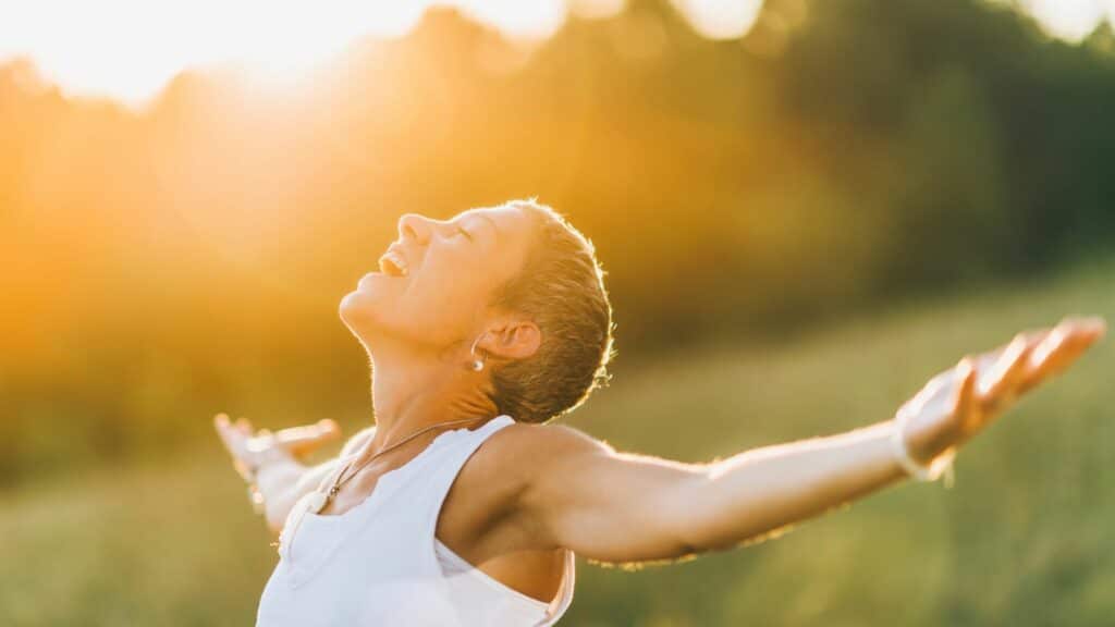 Imagem de uma mulher de cabelos curtos, usando uma camiseta regata na cor branca. Ela está de braços abertos, feliz, olhando para cima e recebendo a energia que vem do sol, simbolizando o conceito de luz interior.

