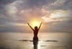 Imagem da silhueta de uma mulher de cabelos curtos, dentro da água do mar. Ela está de braços abertos, feliz, olhando para cima e recebendo a energia que vem do sol, simbolizando o conceito de luz interior.