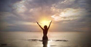 Imagem da silhueta de uma mulher de cabelos curtos, dentro da água do mar. Ela está de braços abertos, feliz, olhando para cima e recebendo a energia que vem do sol, simbolizando o conceito de luz interior.